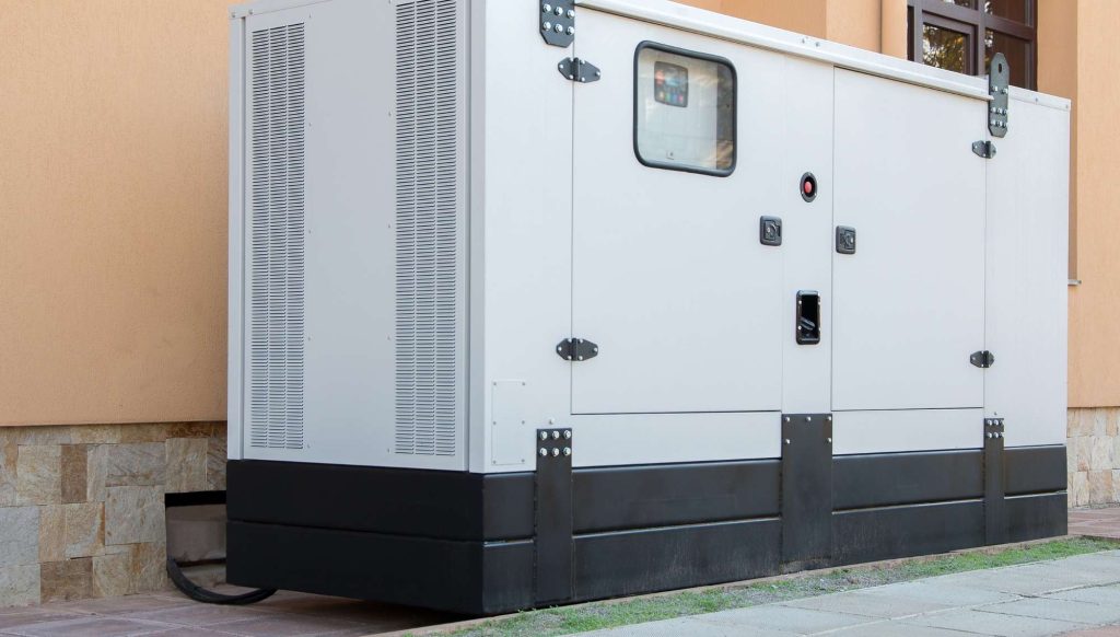 A large white industrial generator with ventilation panels stands next to a building with tan walls and a stone base in AZ. A window and door are visible on the generator’s side, with cables running out—perfect for an electrician Maricopa County might call upon.