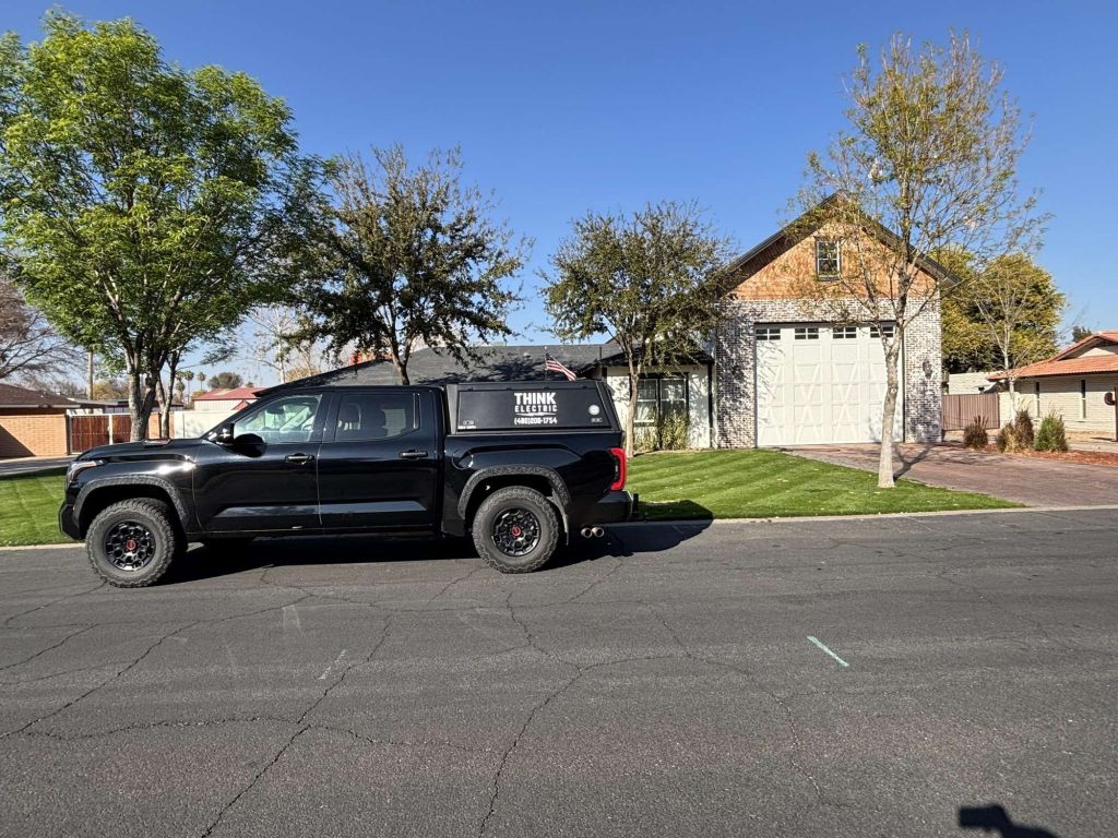 A black pickup truck with “THINK” and a phone number on the side, advertising an electrician Maricopa County, AZ, is parked on a sunny residential street in front of a house with a well-kept lawn, trees, and a large garage.