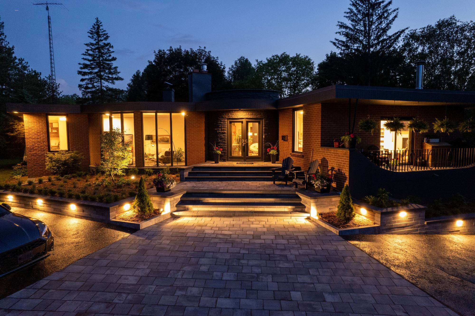 A modern house at dusk with warm exterior lights illuminating the front steps, garden, and driveway, surrounded by trees and greenery.