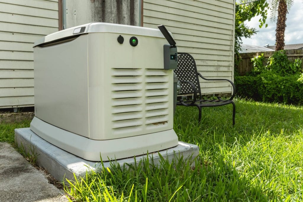 A standby generator sits on a concrete pad next to a house with beige siding and a black metal bench on grass nearby. The yard is bordered by a wooden fence and trees.
