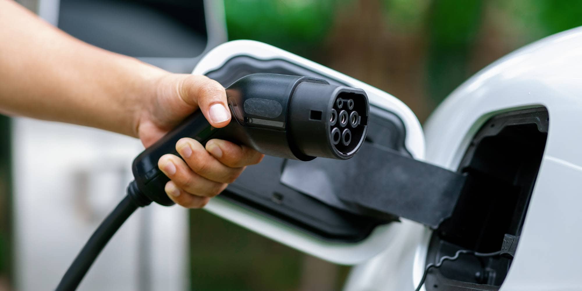 A person’s hand holds an electric car charging plug, preparing to connect it to the charging port of a white vehicle. The background is green and blurred, suggesting an outdoor setting.
