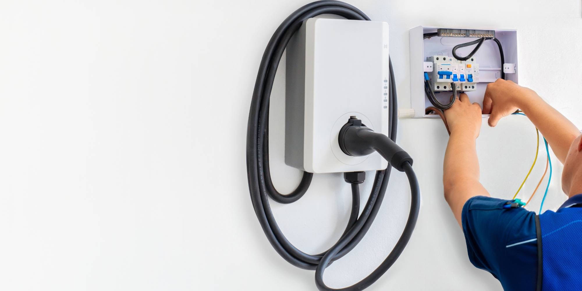 A person in a blue shirt installs or repairs the wiring of an electric vehicle (EV) charging station mounted on a white wall. The charging cable is coiled neatly around the unit.
