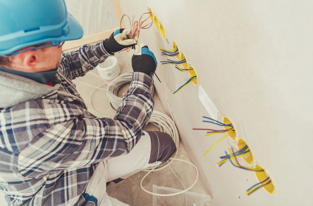 An electrician Maricopa County, AZ, wearing a blue hard hat and gloves, is installing electrical wires into wall sockets, working on a partially finished wall with exposed wiring and cables.