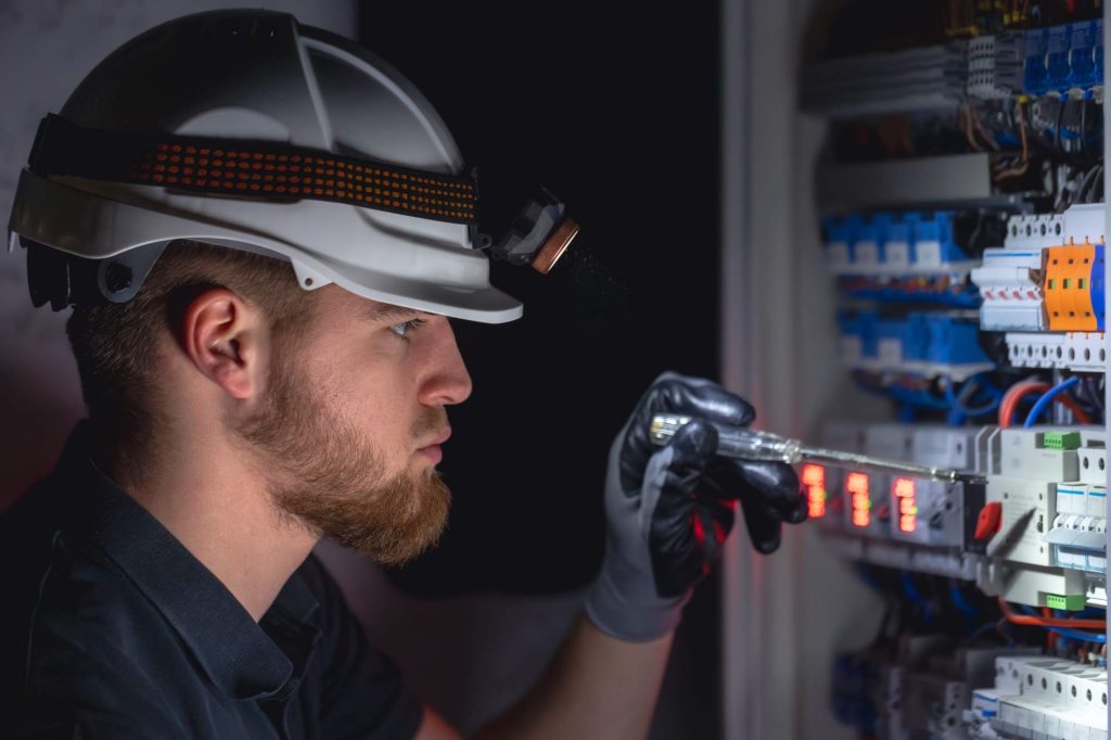 A man wearing a hard hat and gloves uses a flashlight to inspect or work on an electrical panel filled with various wires and switches.