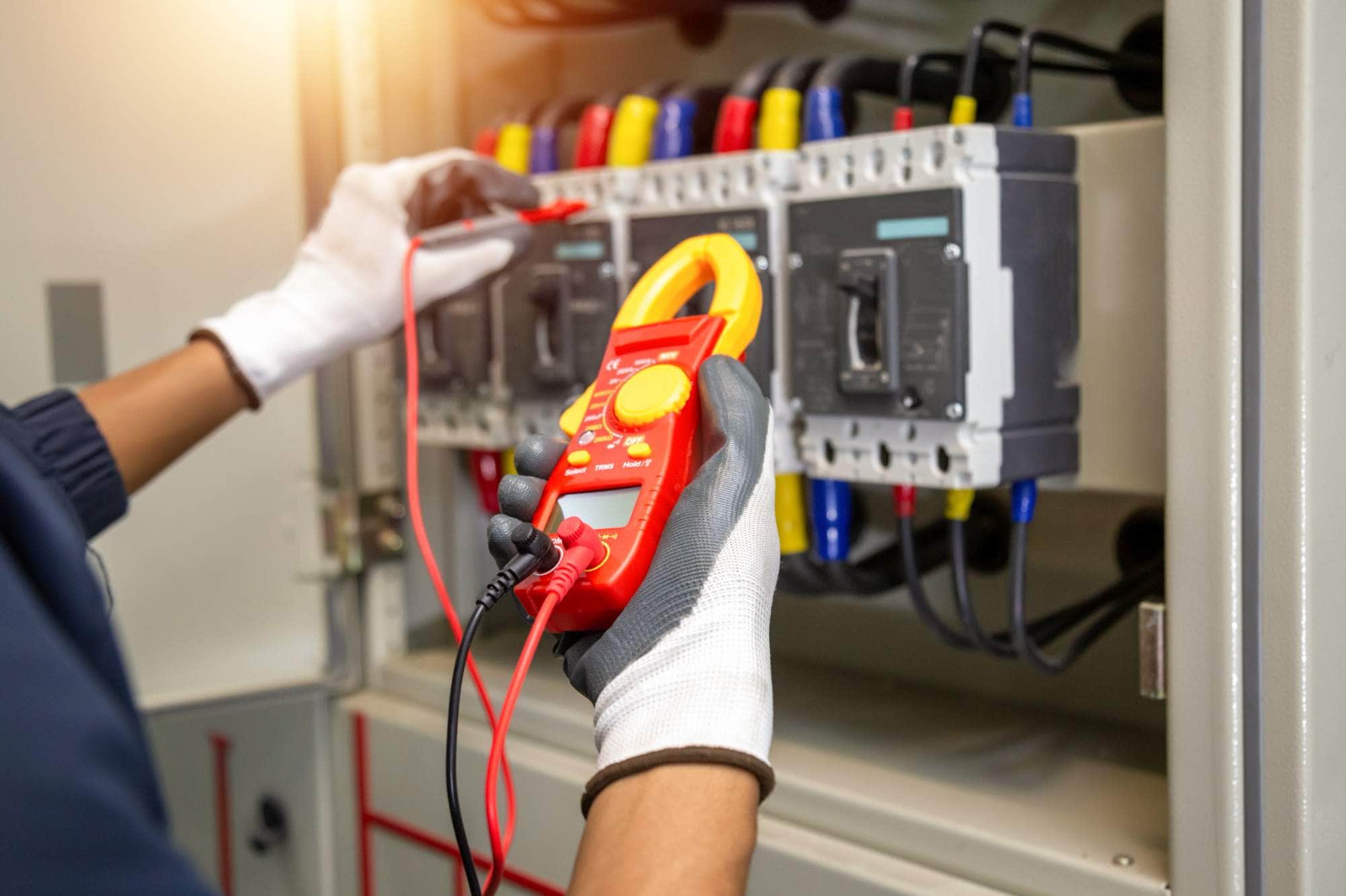 A person wearing white gloves uses a digital multimeter to check electrical parameters inside an industrial control panel with multiple colored wires and circuit breakers.