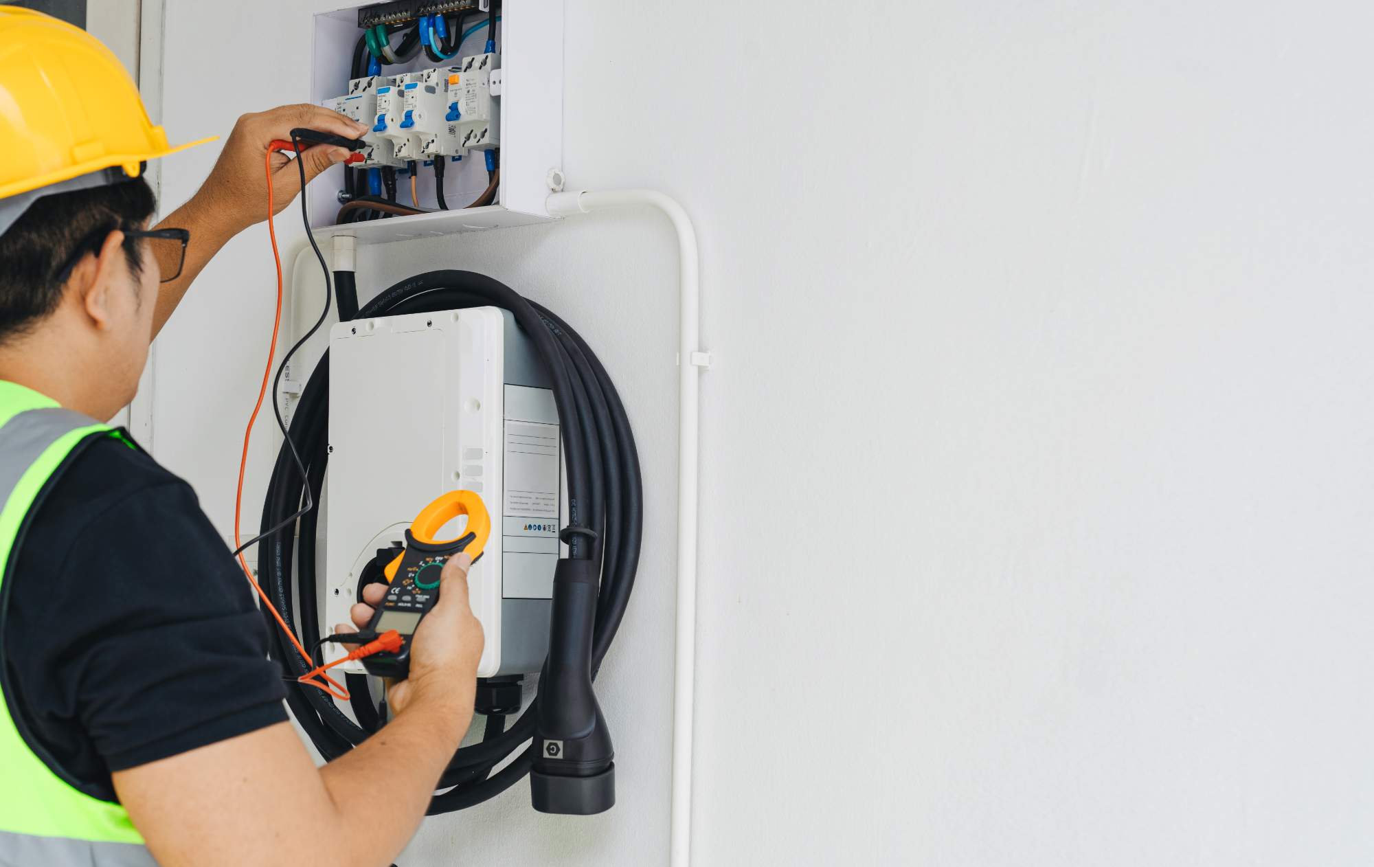 An electrician wearing a yellow hard hat and safety vest uses a multimeter to check an electrical panel mounted on a white wall, with cables and a charging device visible nearby.