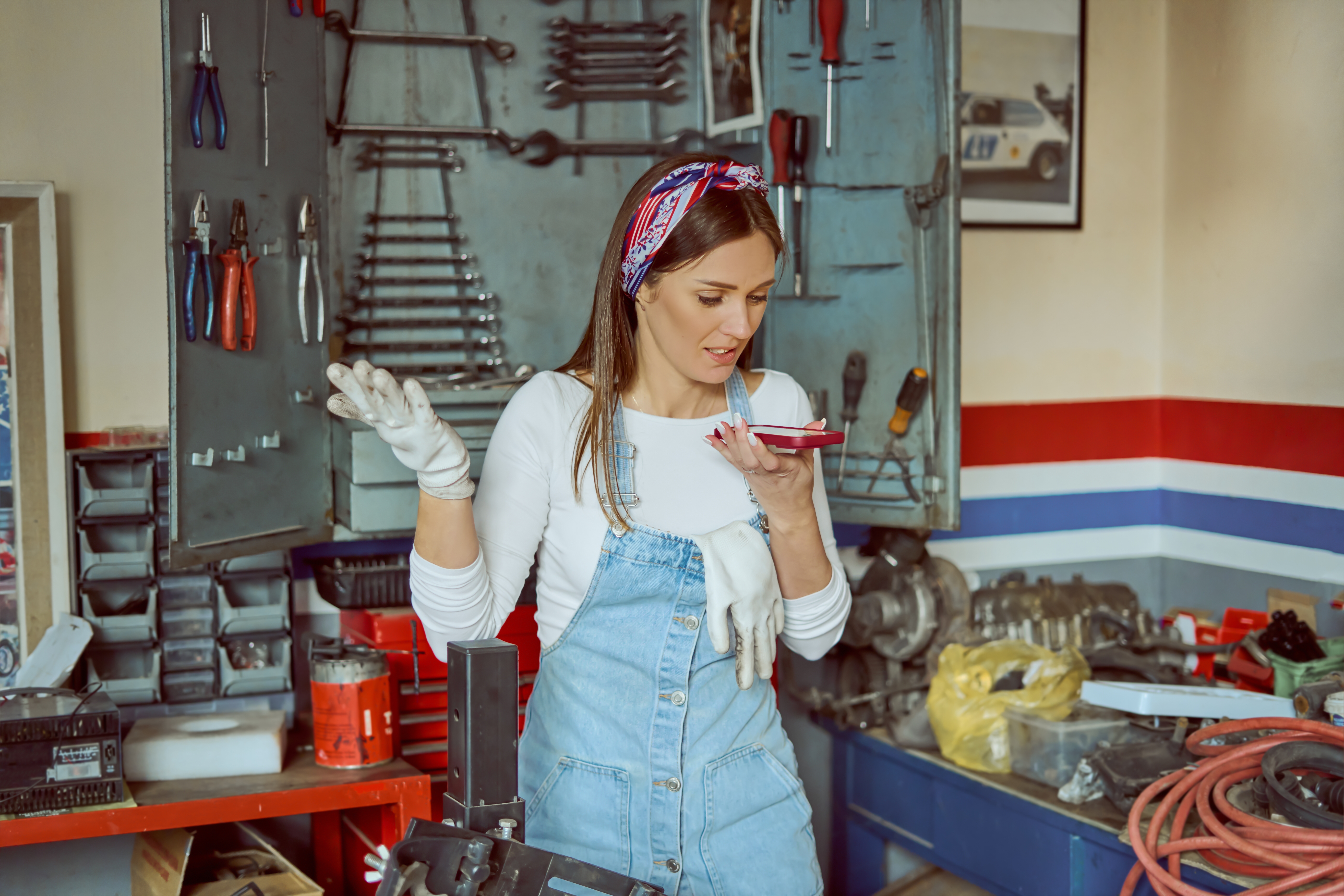 Woman in denim overalls and work gloves on the phone in a cluttered garage with a tool wall, showing the need for a garage cabinet system to organize